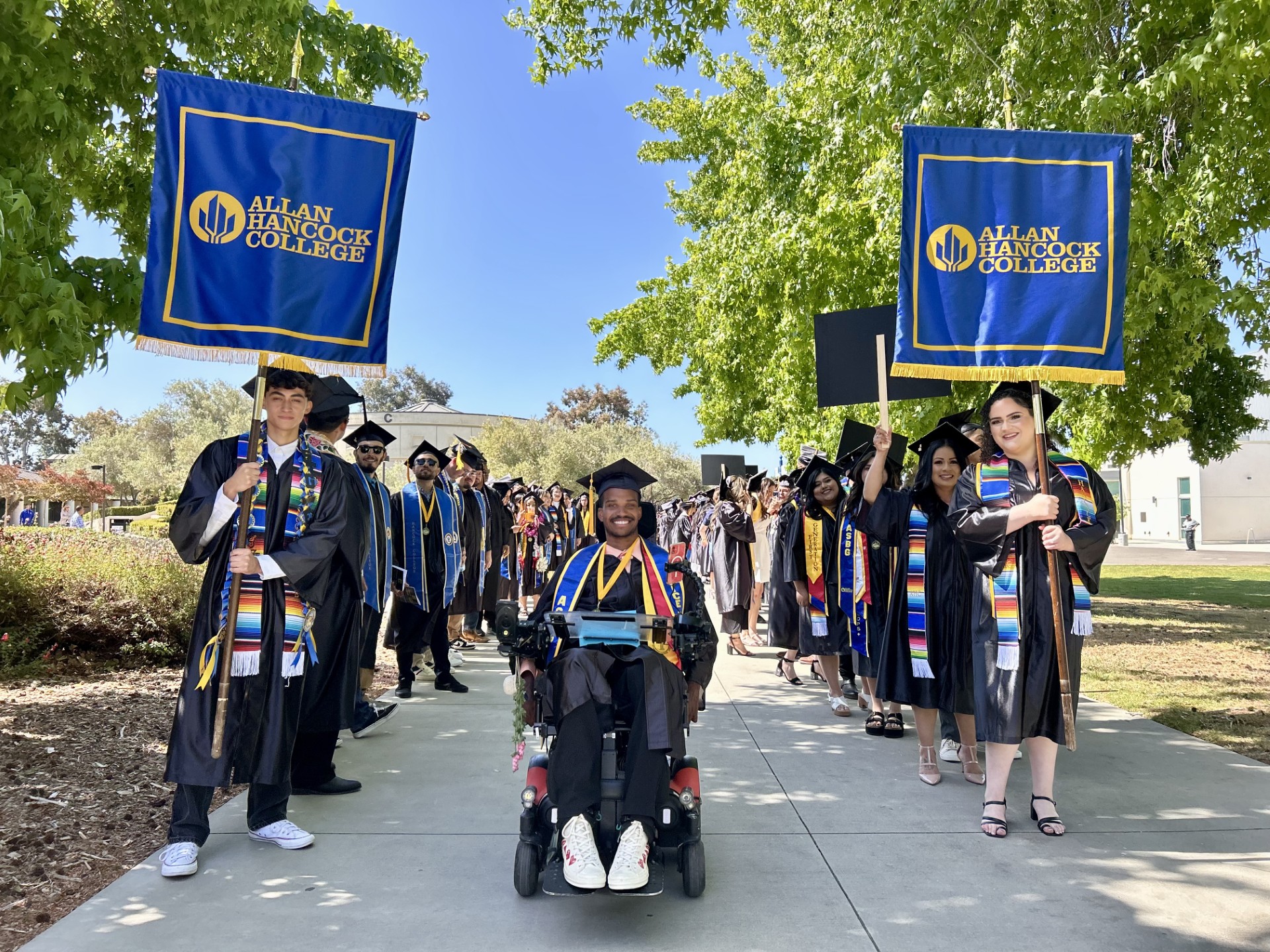 Graduates from Allan Hancock College in caps and gowns smile as they line up in two rows outdoors under leafy trees. Two students at the front hold banners with the college logo, while a graduate in a wheelchair leads the procession down a sidewalk on a sunny day.